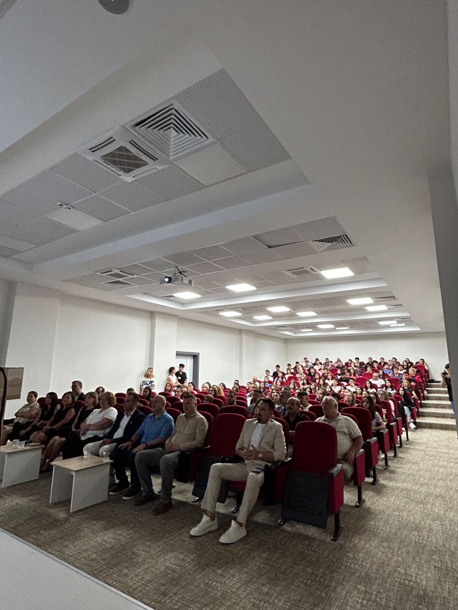 Bu kare bir konferans salonunda çekilmiş. Salon geniş, beyaz duvarlı ve tavanında kare şeklinde  aydınlatmalar var.Kırmızı koltuk sıraları yukarıya doğru yükseliyor. Ön sıralarda takım elbiseli ve sade giyimli öğretim elemanları oturuyor; çoğu ciddi ama dikkatli bir şekilde sahneye doğru bakıyor.  Arka sıralar öğrencilerle dolu, Salonda projektör cihazı tavandan sarkıyor ve bir  sunum yapılmakta. Ortam resmi ama samimi bir havada