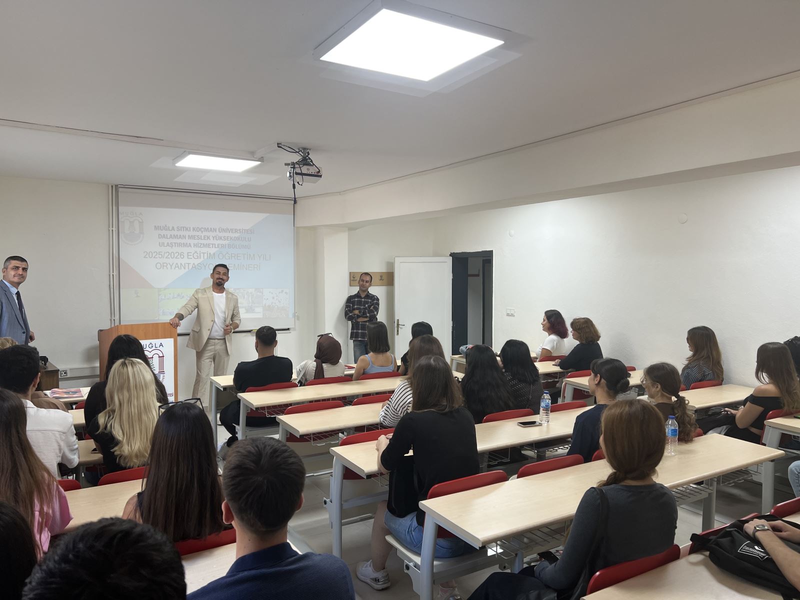 Bu fotoğraf bir sınıf ortamında çekilmiş.Ön planda sıralarda oturan genç öğrenciler var; çoğu kadın, birkaçı erkek. Hepsi tahtaya veya konuşmacıya dikkatle bakıyor.Önde Bölüm Başkanı konuşma yapıyor; açık renk takım giymiş ve el hareketleriyle anlatımını destekliyor.Arka planda bir projeksiyon perdesi var; üzerinde şu yazı okunabiliyor:“Muğla Sıtkı Koçman Üniversitesi, Dalaman Meslek Yüksekokulu, Ulaştırma Hizmetleri Bölümü, 2025/2026 Eğitim Öğretim Yılı Oryantasyon Semineri.”  Konuşmacının yanında bölümdeki diğer iki öğretim elemanı var var; biri takım elbiseli, diğeri gömlekli. Sınıf ortamı düzenli, aydınlık ve sıcak bir atmosfer taşıyor.