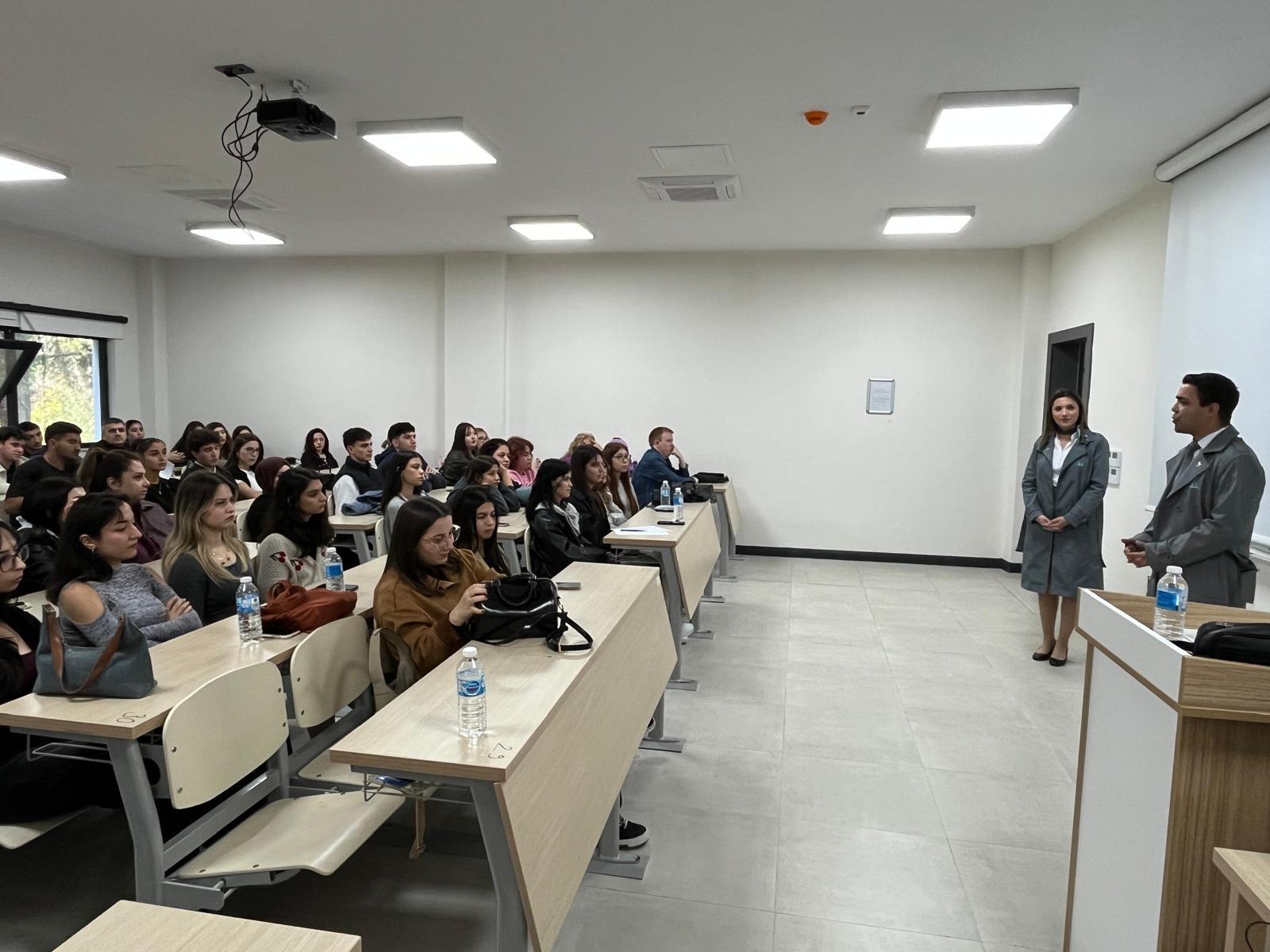 This wide-angle photo captures a bright classroom or lecture hall. On the left side, approximately 30 students are seated at desks, facing the front. The seating consists of horizontal, wooden desks and white, metal-framed chairs. On the right side, in front of a podium, the male and female speakers in the grey/blue trench coats from the first photo are standing, addressing the students. The walls are white, and the ceiling is lit with square LED lights.