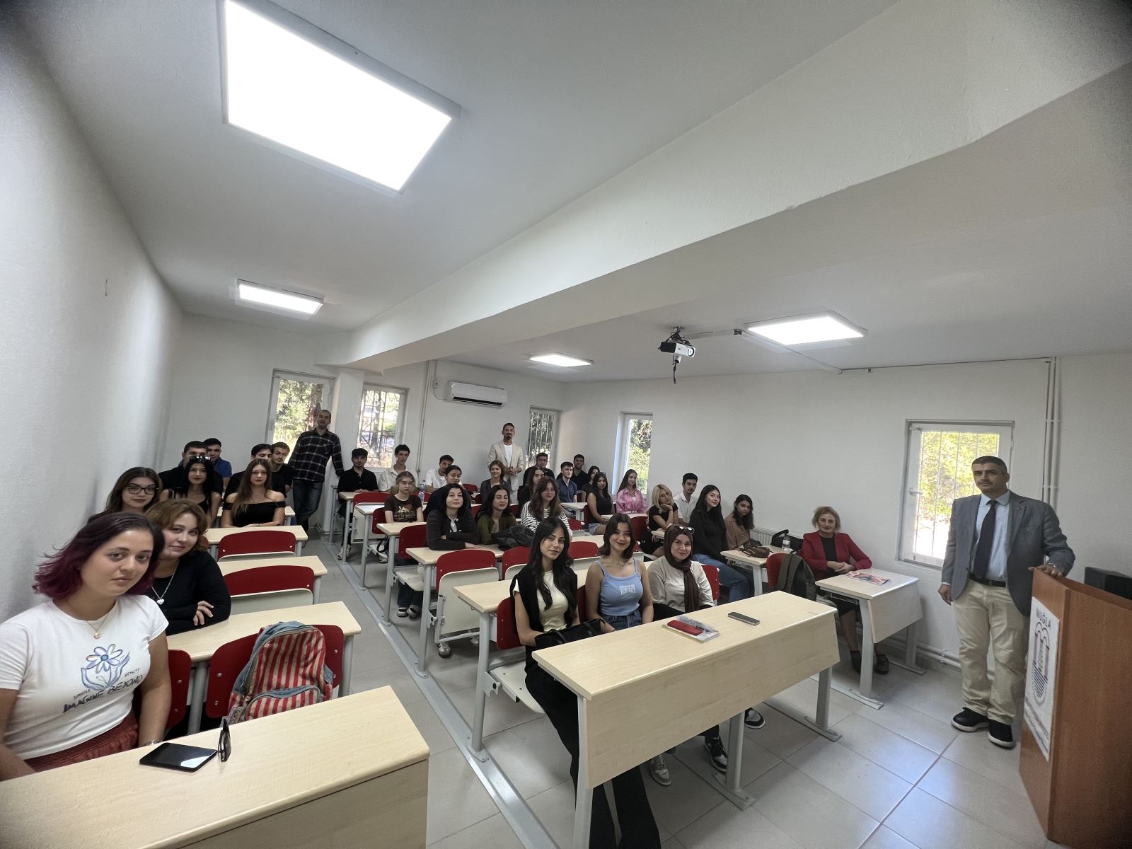 Bu fotoğraf bir sınıf ortamında çekilmiş.Öğrenciler sıralarda oturuyor öğrencilerin araların üç erkek bölüm öğretim elemanları ayakta duruyor Hepsi sınıfın ön kısmında bulunan fotoğrafı çeken kişinin objektifine gülümseyerek bakıyor