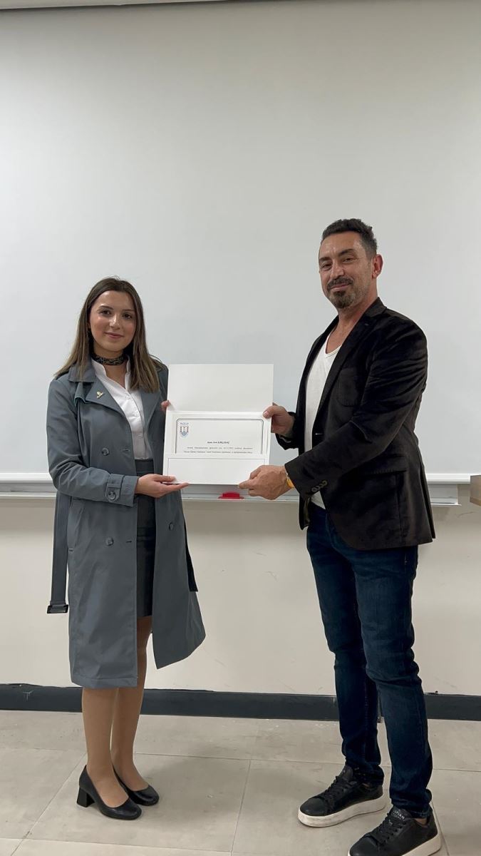 This vertical photograph also shows two people standing in front of a white screen. The woman on the left, wearing the grey trench coat, is presenting a certificate to, or holding it with,