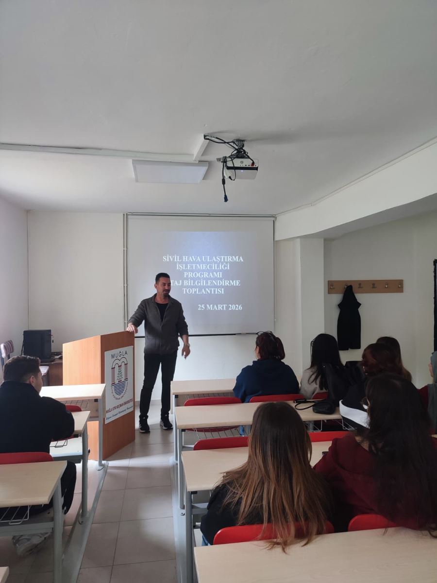 The photograph captures an information meeting taking place in a classroom or seminar hall.  • The Presenter: A middle-aged man with dark hair and a beard stands at the front. He is wearing a grey cardigan over a black t-shirt, resting one hand on a lectern and smiling towards the audience.  • Projection Screen: Behind him, a presentation is projected onto a white wall. The Turkish text translates to "CIVIL AIR TRANSPORT MANAGEMENT PROGRAM INTERNSHIP INFORMATION MEETING" with the date "MARCH 25, 2026" displayed below.  • Lectern: To the man's left is a wooden lectern featuring the logo and name of Muğla Sıtkı Koçman University.  • Foreground: In the lower part of the image, classroom desks and chairs are visible. Two female students are seated in the front row with their backs to the camera. The desks have light-colored wooden tops and metal frames, paired with bright red chairs.  • Setting: The room is well-lit, with a projector mounted on the ceiling. On the right wall, there is a coat rack with a black jacket hanging on it.