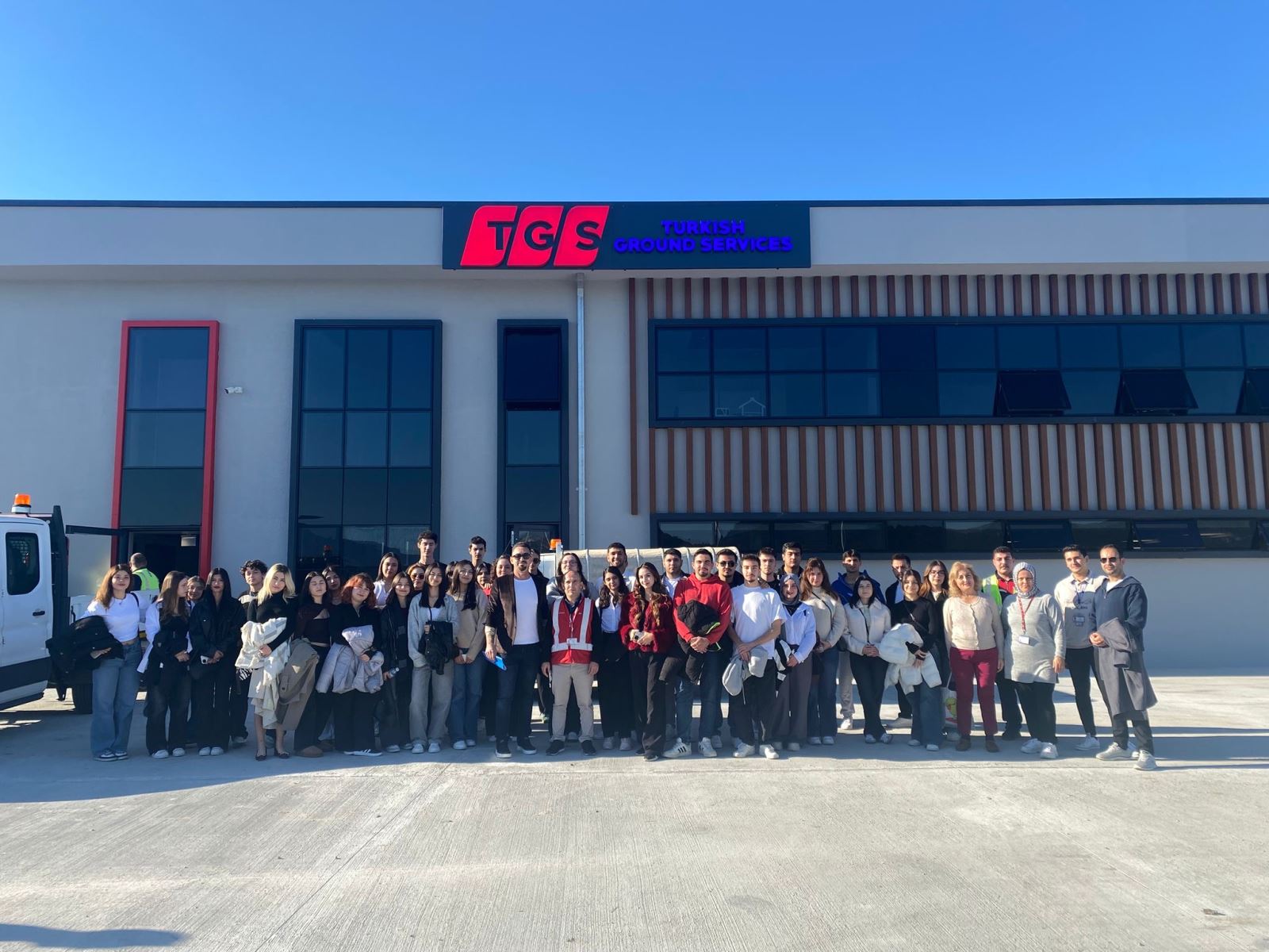 Bu karede, kalabalık öğrenci grubu modern ve gri bir binanın önünde toplu halde poz veriyor. Binanın üst kısmında kırmızı logolu "TGS Turkish Ground Services" tabelası yer alıyor. Binanın pencereleri geniş ve siyah çerçeveli; sağ tarafta ise dikey ahşap çıtalarla yapılmış dekoratif bir dış cephe bölümü görülüyor. Grubun ortasında kırmızı yelekli bir yetkili bulunuyor. Hava oldukça güneşli ve gökyüzü bulutsuz.